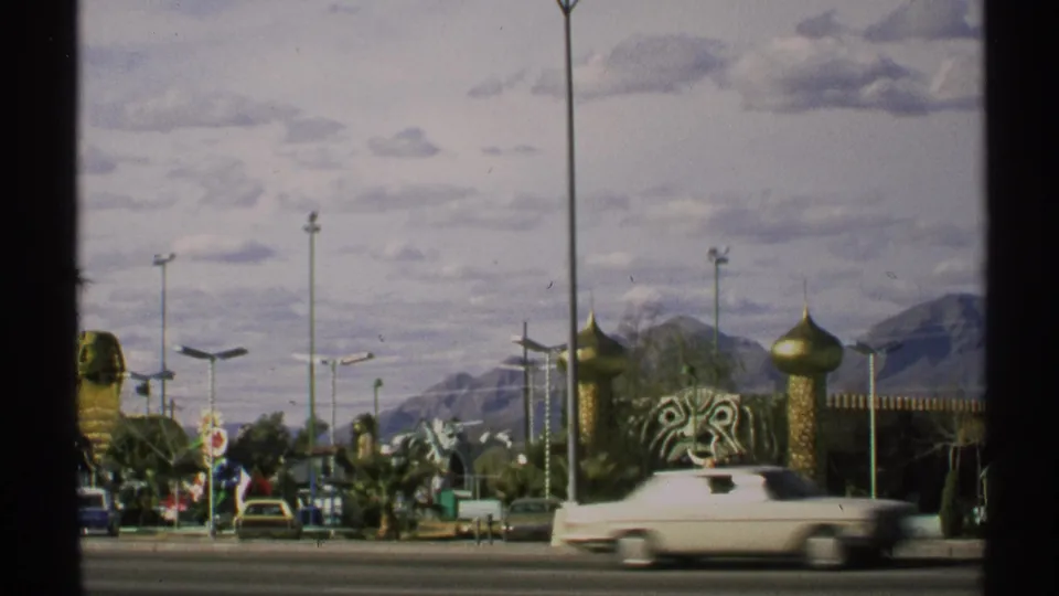 Moving 1972 Arizona view of roadside landmark structures, a passing white car, lamp posts, desert plantings, and mountains under a cloudy sky.