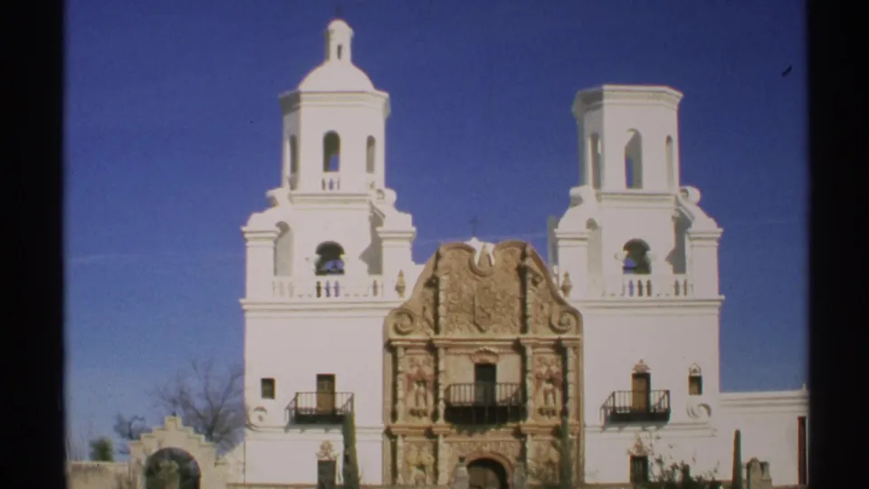 1972 Arizona frame of a white Spanish colonial-style church exterior with twin bell towers, ornate stonework, balconies, and blue sky.