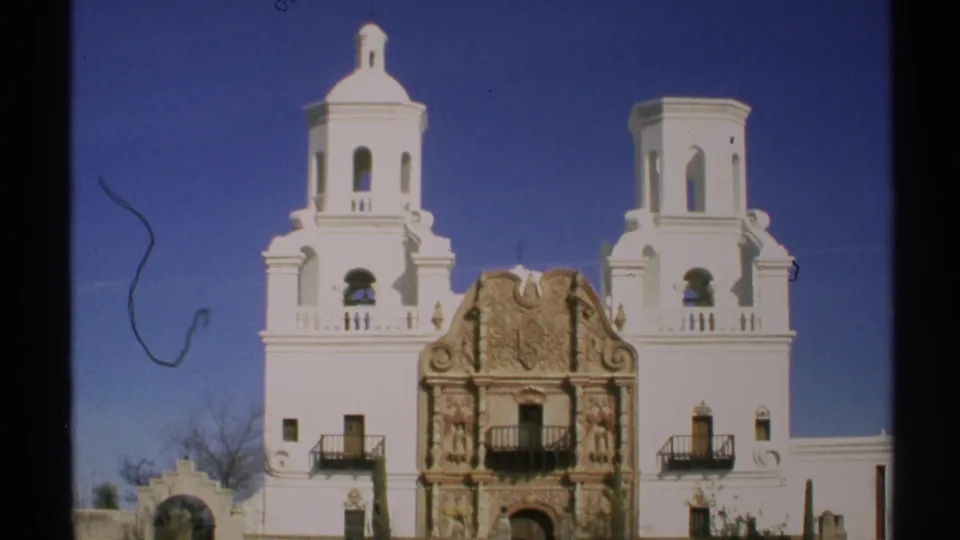 1972 Arizona home-movie frame showing the white twin-tower church facade, carved stone entrance, small balconies, and bright blue desert sky.
