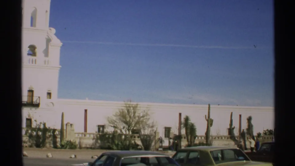 Wider Arizona street view shows the church wall, cacti, desert landscaping, and parked early 1970s cars in the foreground.