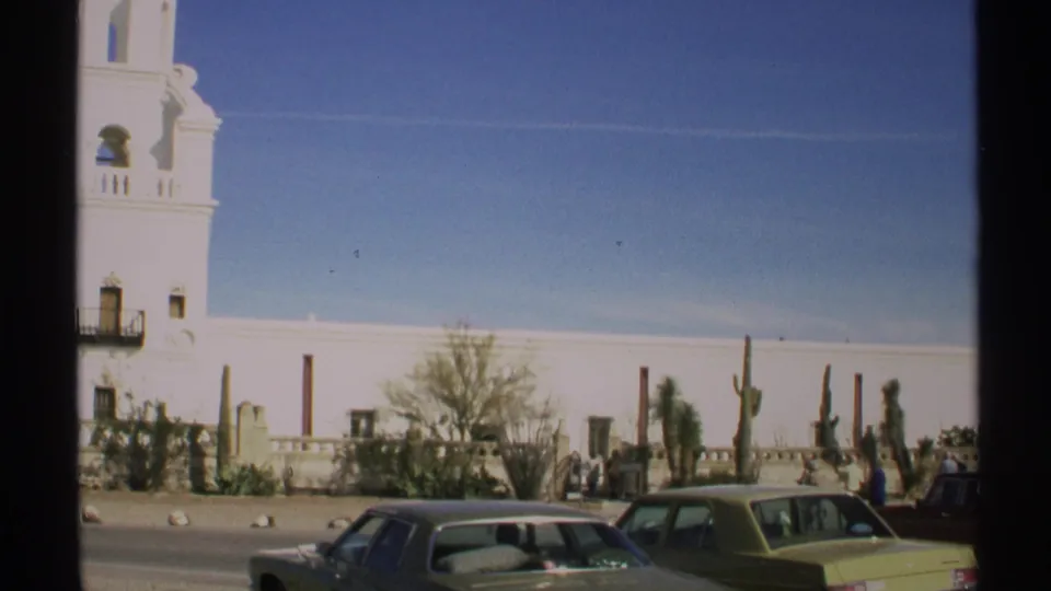 The final 1972 Arizona frame widens to show church grounds, desert plants, parked sedans, and small visible visitors near the roadside.