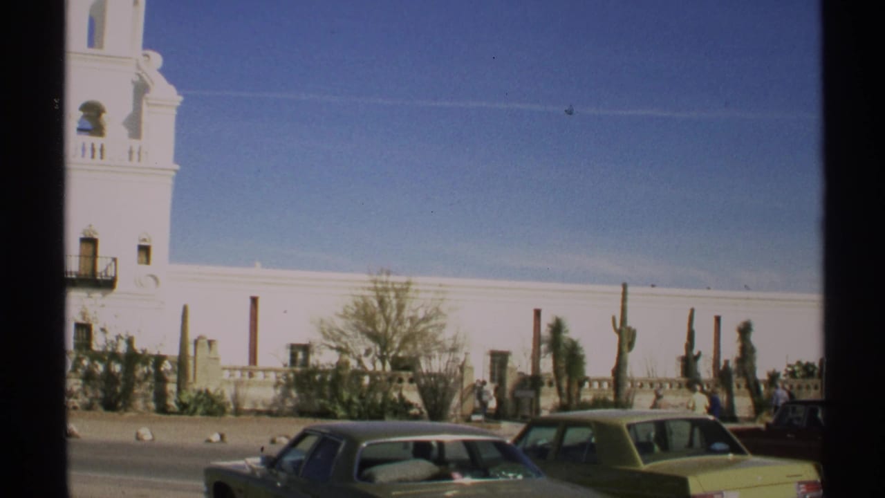 White adobe-style building with tower, parked cars, cactus, and pedestrians along a sunny street.