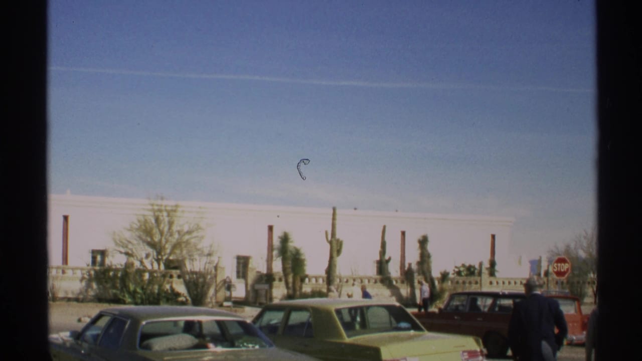 Parked cars and pedestrians in front of a long white building with cactus and a stop sign.
