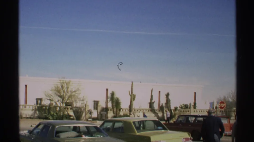 1972 Arizona street view with period cars, visitors, cactus plantings, a stop sign, and a long white adobe-style facade.