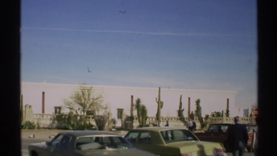 1972 Arizona 8mm frame showing a low white building, period parked cars, cactus, pedestrians, and a red stop sign.