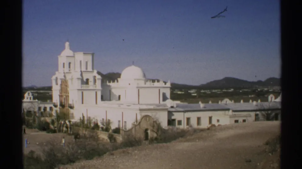 1972 Arizona frame showing a white domed adobe-style building, bell tower, desert ground, nearby structures, and distant hills.