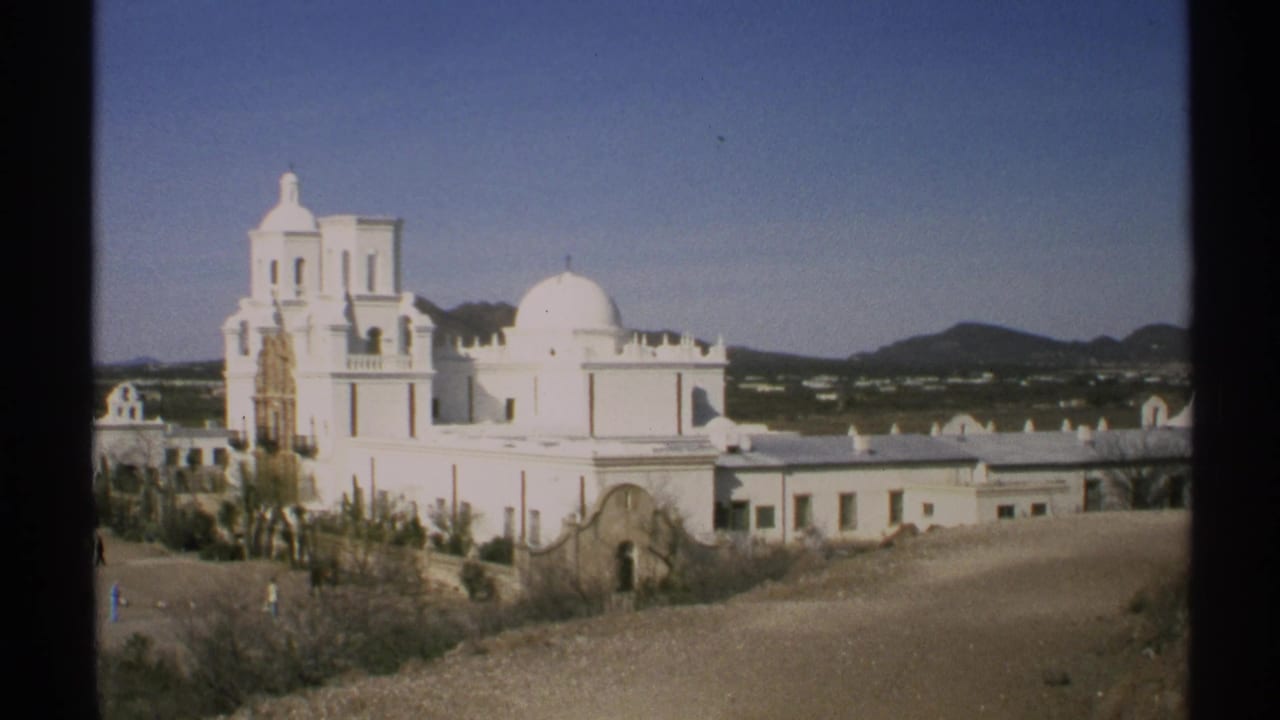 1972 Arizona 8mm frame showing a white church complex with dome, bell tower, low shrubs, and distant mountains.