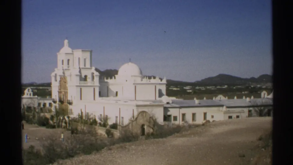1972 Arizona 8mm frame showing a white church complex with dome, bell tower, low shrubs, and distant mountains.