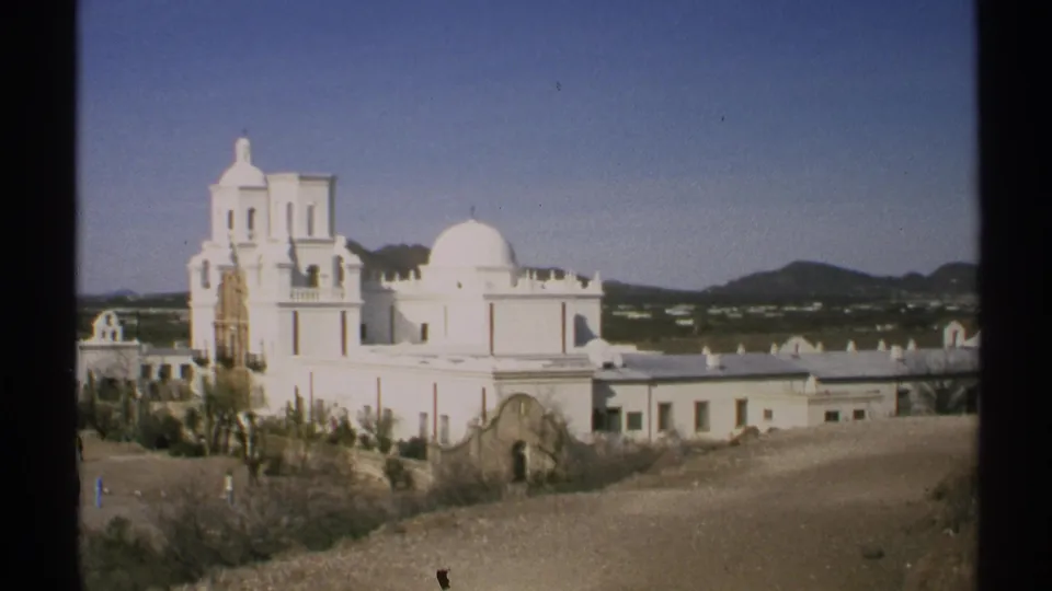 1972 Arizona frame holding on the white church, dome, bell tower, foreground brush, and clear blue sky.