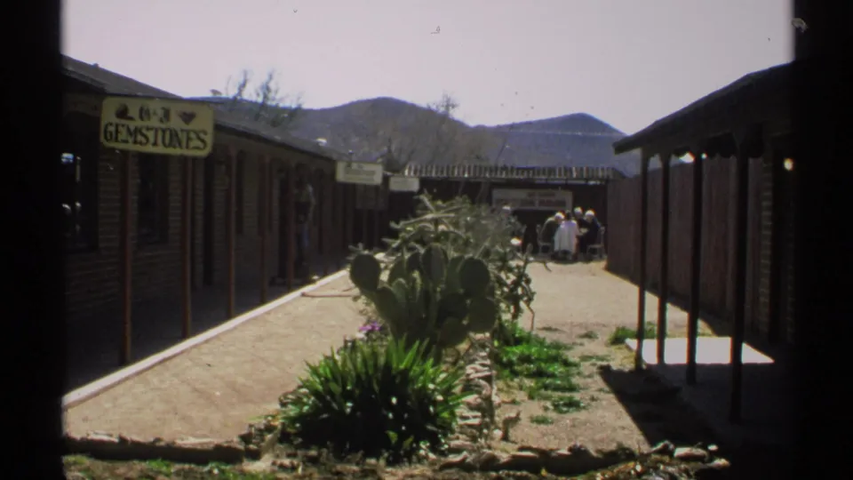 1972 Arizona 8mm frame showing a desert shop corridor, GEMSTONES sign, cactus plantings, distant mountains, and small visitor figures.