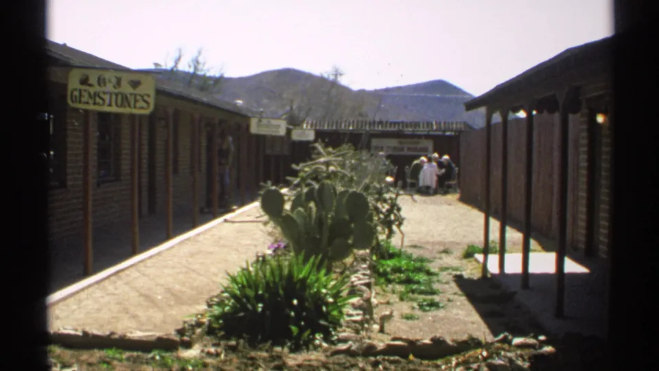1972 Arizona home-movie frame of a gemstone shop exterior with cactus landscaping, rustic buildings, mountains, and visitors at the far end.