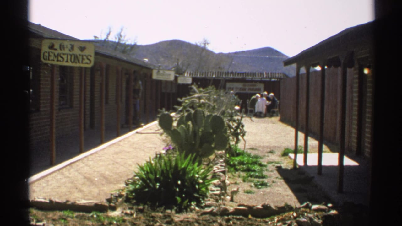 Desert shop courtyard with GEMSTONES sign, cacti, brick storefronts, distant visitors, and mountains.