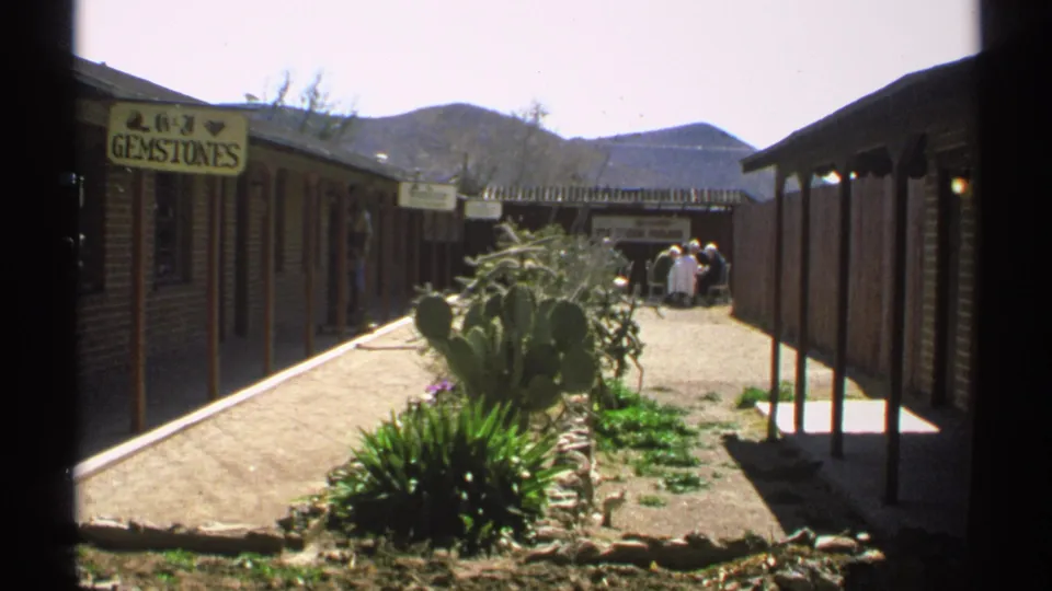 1972 Arizona 8mm frame showing a desert gemstone shop courtyard with cacti, brick storefronts, visitors in the distance, and mountains beyond.