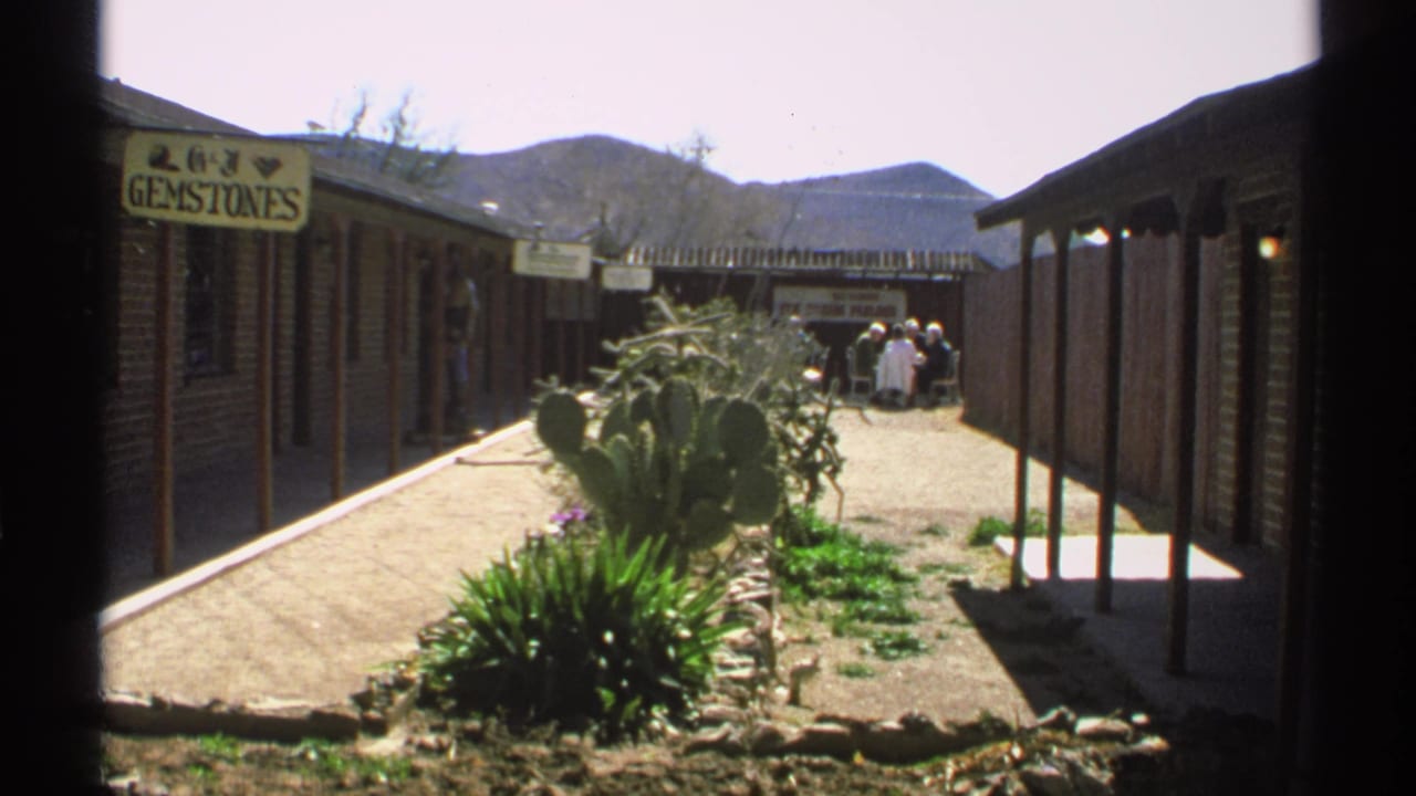 Similar courtyard view with gemstone shop sign, cactus bed, brick buildings, people, and mountains.