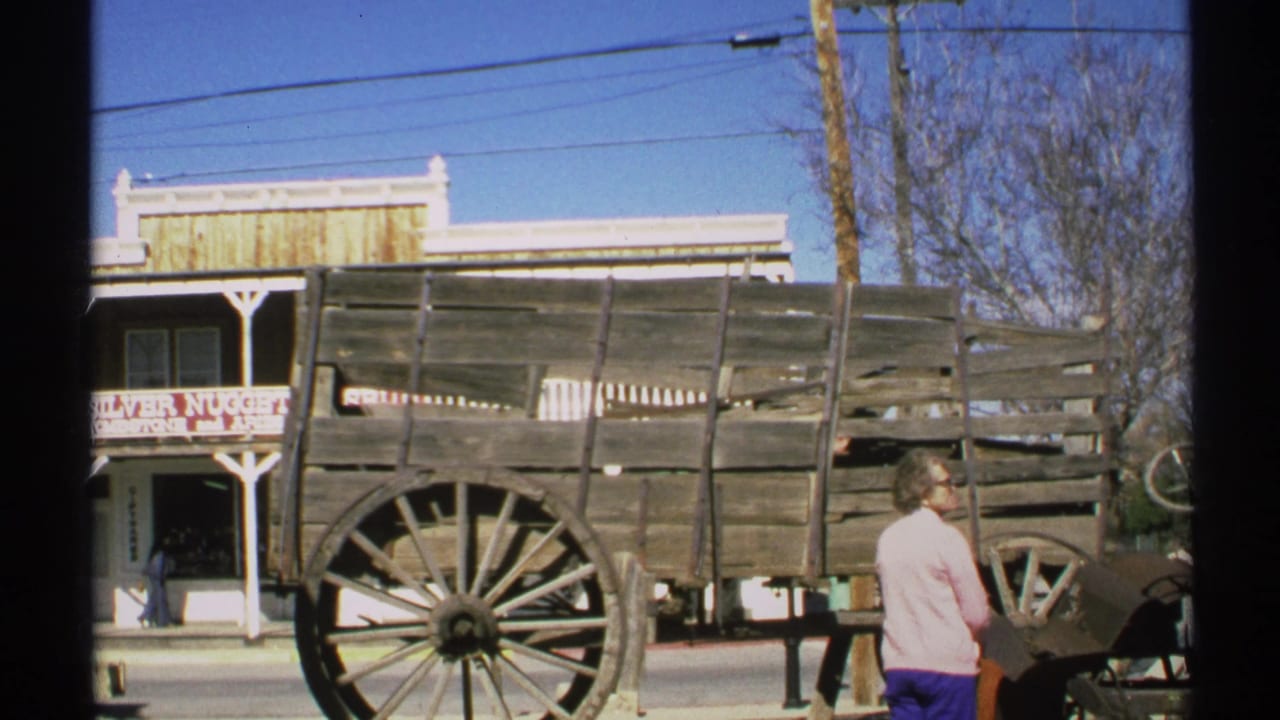1972 Arizona home-movie frame showing an older woman near a weathered wooden wagon with a Silver Nugget sign behind it.