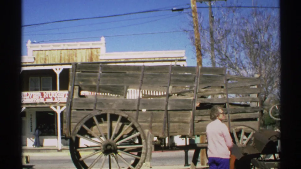 1972 Arizona home-movie frame showing an older woman near a weathered wooden wagon with a Silver Nugget sign behind it.