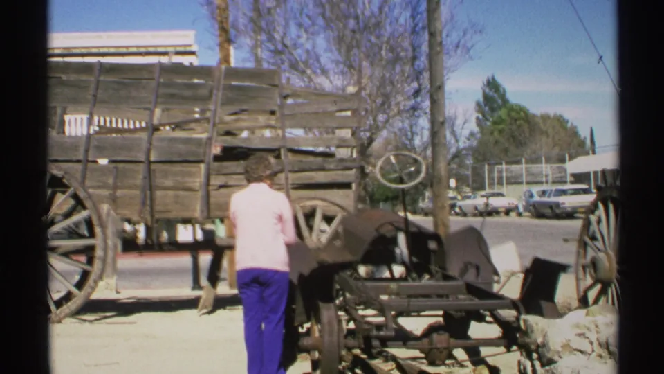 1972 Arizona home-movie frame of a woman near wagon wheels and street display equipment, with parked cars in the background.