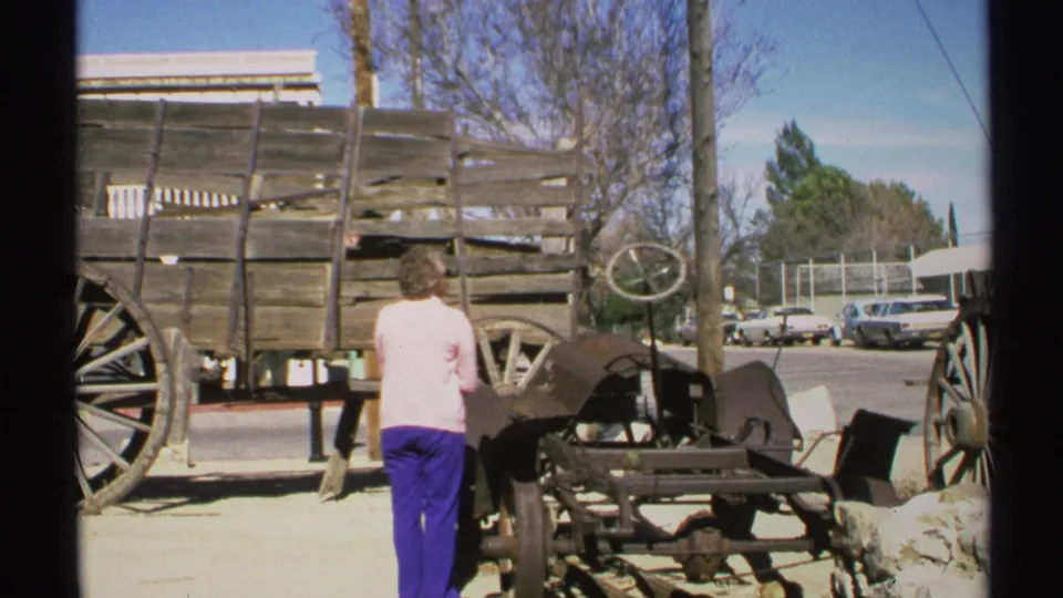 1972 Arizona frame showing the woman beside a wooden wagon and metal street display, with mid-century cars beyond.