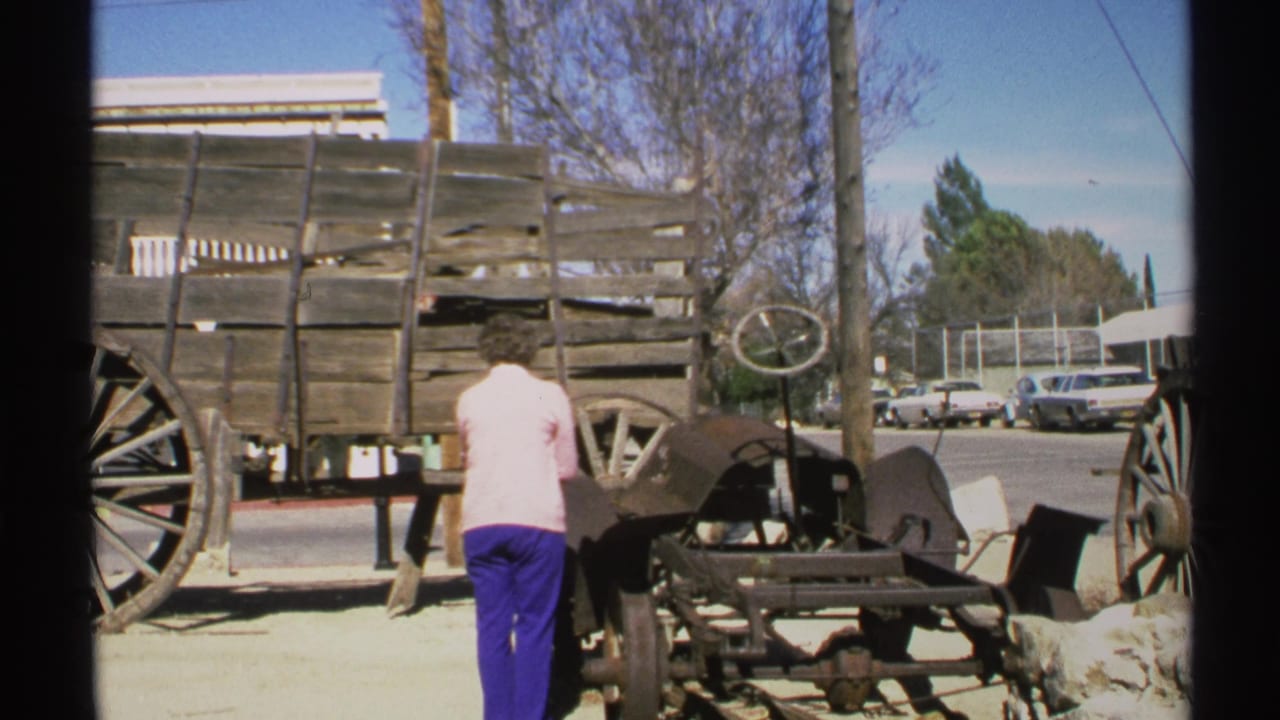 In 1972 Arizona home-movie footage, a woman in blue pants and a pink top stands near old wagon parts beside parked cars and buildings.