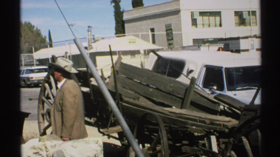 A man in a cowboy hat and brown jacket stands near weathered wagon parts in a 1972 Arizona outdoor roadside setting.