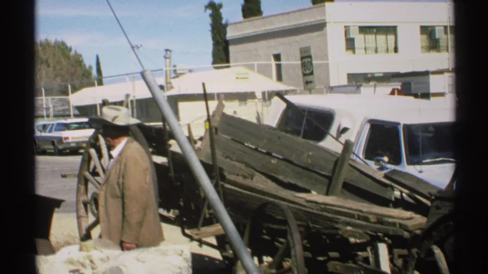 A 1972 Arizona home-movie frame shows a man beside weathered wagon parts, with parked vehicles, buildings, and a visible road sign behind.