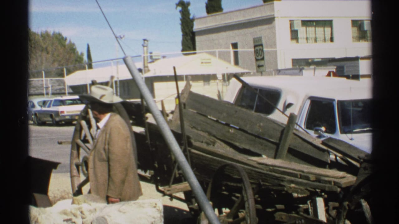 In Arizona 1972 footage, a man in a light suit and wide-brimmed hat stands beside an old wooden wagon near cars, a building, and a Route 80 sign.