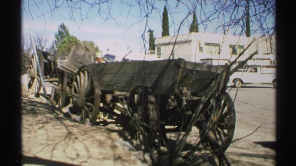 Arizona 1972 home-movie frame showing a weathered wooden wagon near a roadside building, parked vehicles, bare branches, and strong sun.