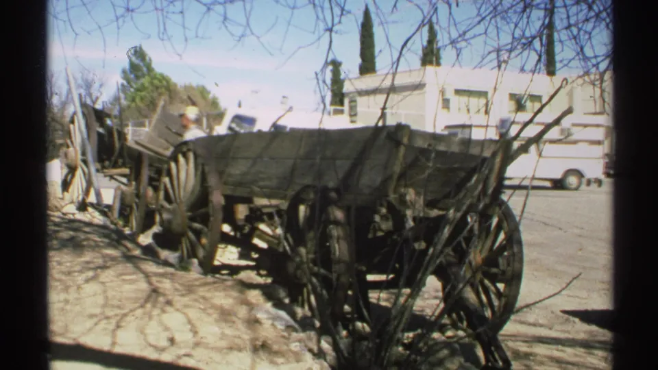 A 1972 Arizona roadside wagon display fills the frame, with a partly visible man behind it, parked vehicles, a pale building, and branch shadows.