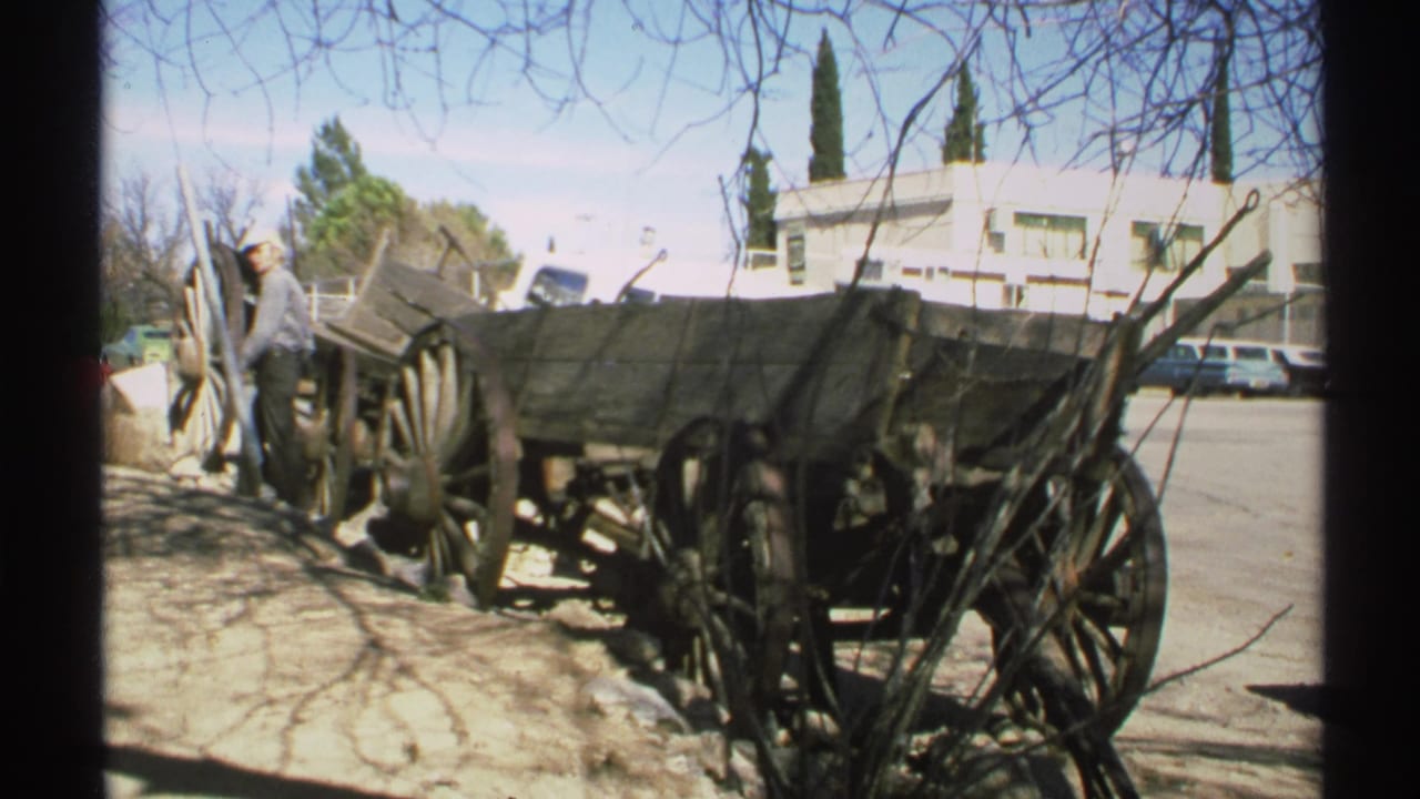 1972 Arizona home-movie frame showing an old wooden wagon display near a street, with a man nearby and low buildings behind it.