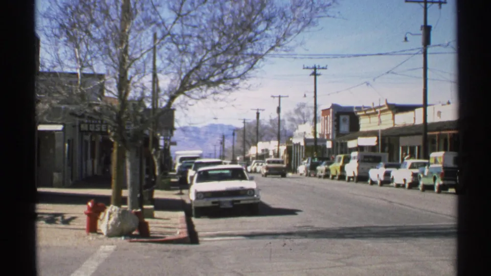 1972 Arizona street view with classic cars parked along storefronts, a red hydrant on the sidewalk, bare trees, and distant mountains.