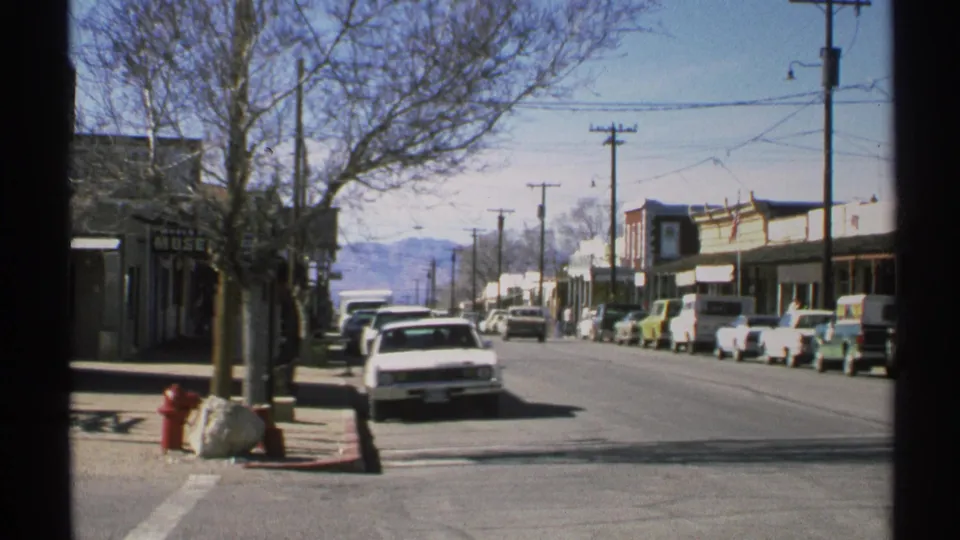 1972 Arizona home-movie view down a small-town street with a white car near the curb, parked vehicles, storefronts, and mountains in the distance.