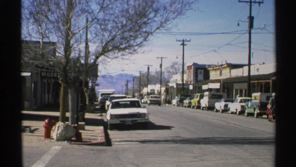 1972 Arizona street scene showing rows of parked cars, storefronts, a red hydrant, bare trees, utility poles, mountains, and a person at frame edge.