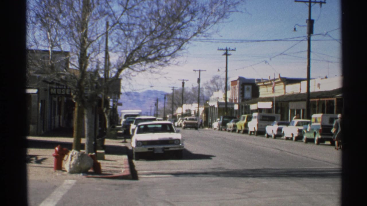 Small-town street with parked cars, storefronts, utility poles, bare trees, and distant mountains.