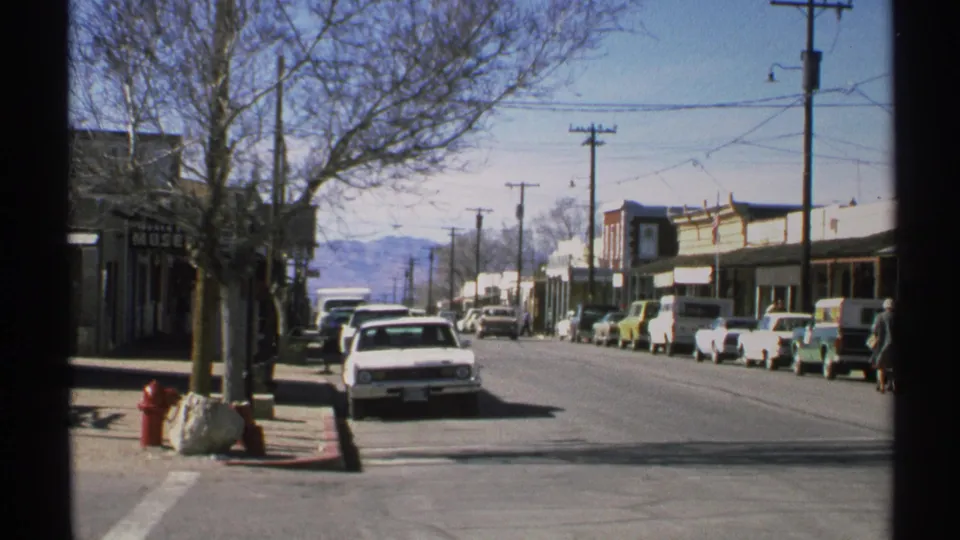 1972 Arizona 8mm view down a sunlit small-town street with classic cars, low storefronts, utility poles, and mountains in the distance.