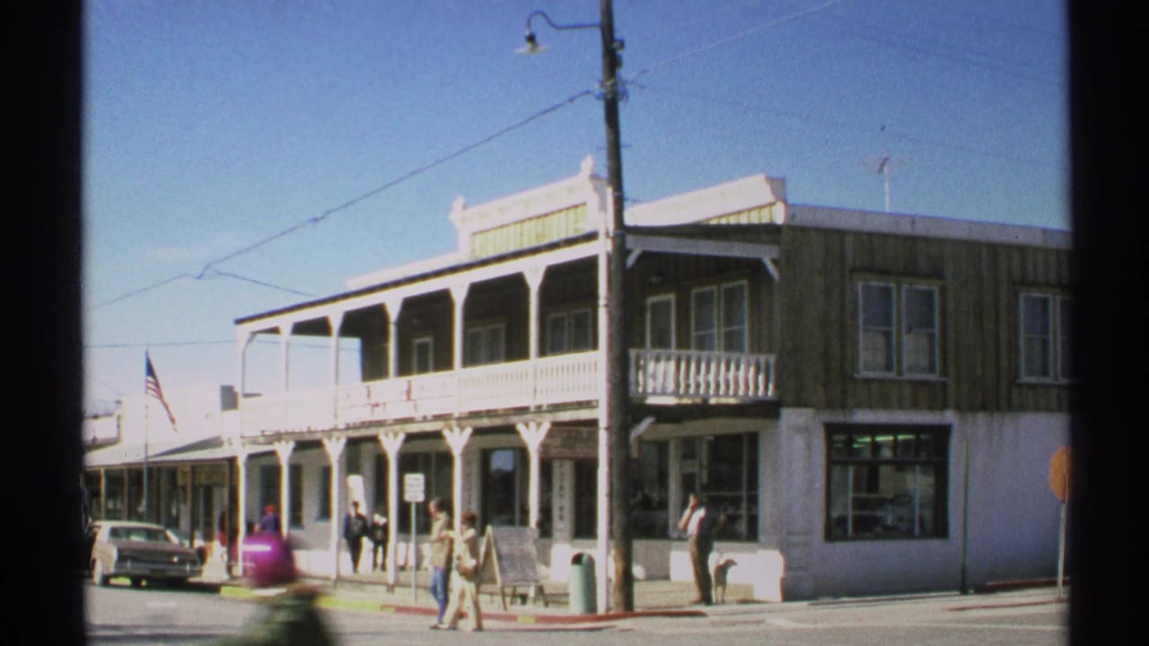 Two-story corner building with wooden porch, pedestrians, parked car, flag, and utility pole.