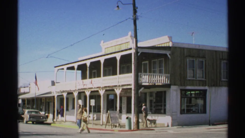 1972 Arizona 8mm frame of a corner storefront with a balcony, people walking on the sidewalk, a parked car, and overhead street wires.