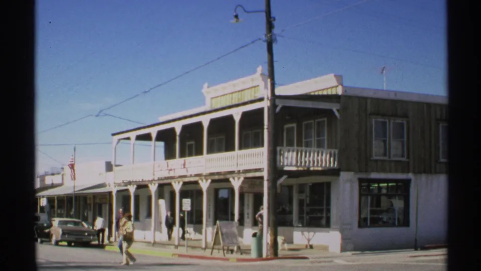 1972 Arizona street-corner view with pedestrians near a wooden-balcony storefront, parked classic car, American flag, and overhead wires.