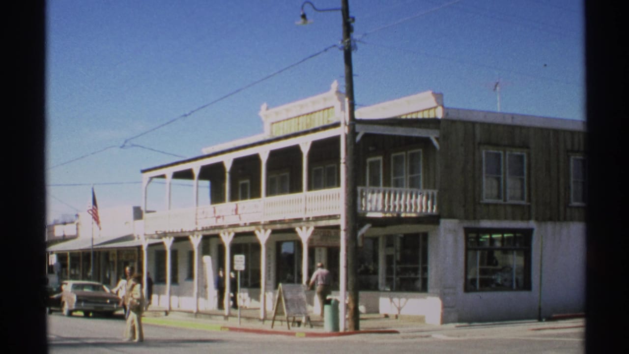 Two-story storefront building with porches, pedestrians, parked cars, a utility pole, and an American flag.