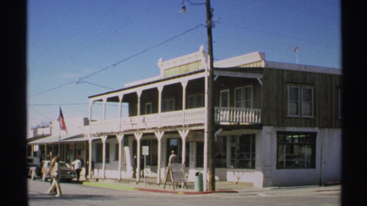 Similar storefront view with pedestrians, parked cars, balcony railings, utility pole, and American flag.
