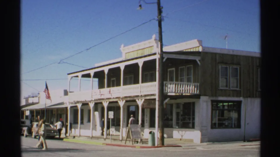 1972 Arizona home-movie frame of a small-town commercial block with porch railings, pedestrians, parked cars, a utility pole, and flag.