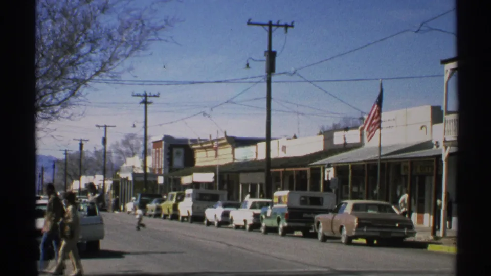 1972 Arizona street scene looking down a commercial block with parked vintage cars, pedestrians, storefront awnings, utility poles, and a flag.