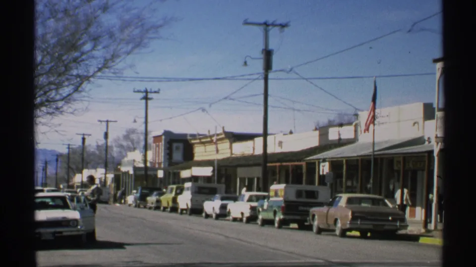 1972 Arizona commercial street view with vintage cars parked along the curb, storefronts under awnings, utility poles, overhead wires, and pedestrians.