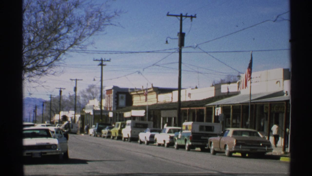 1972 Arizona small-town main street with early 1970s cars, storefronts, overhead wires, pedestrians, and an American flag.