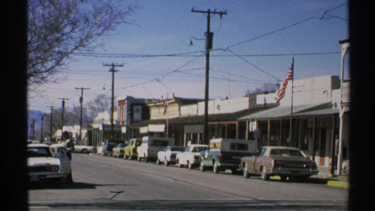 Main street view with classic cars, low storefronts, utility wires, and a flag.