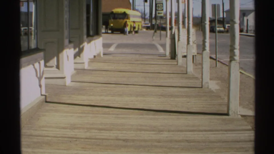 Porch-side view in the stated 1972 Arizona setting, looking down a wooden sidewalk toward a yellow school bus and Route 80 sign.