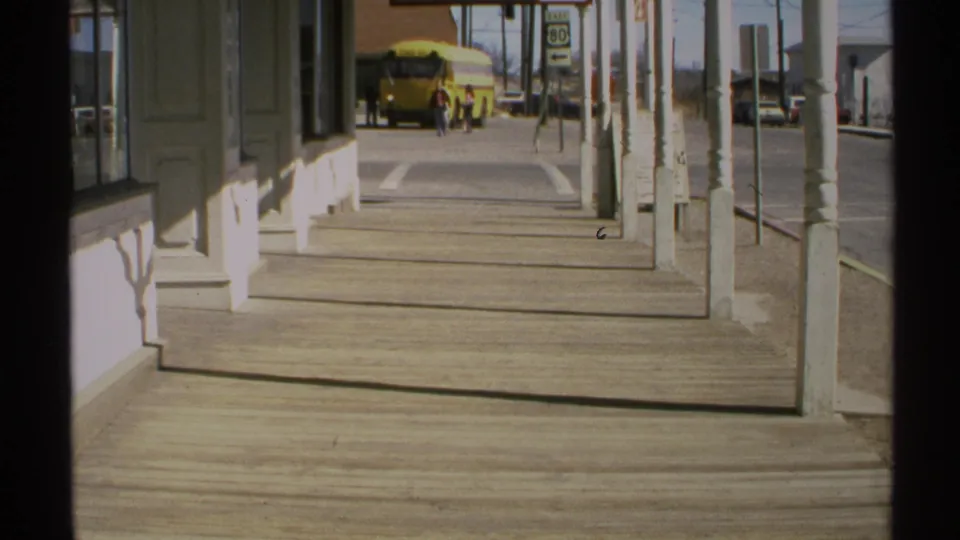 1972 Arizona porch perspective with storefront columns, wooden sidewalk boards, a roadside sign, and a yellow school bus down the street.