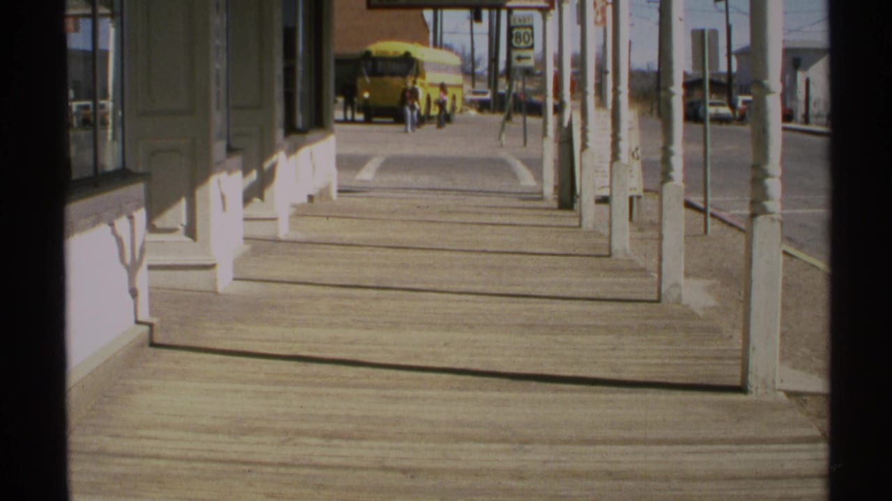 1972 Arizona 8mm frame looking down a covered boardwalk toward a yellow school bus and distant pedestrians on a small-town street.