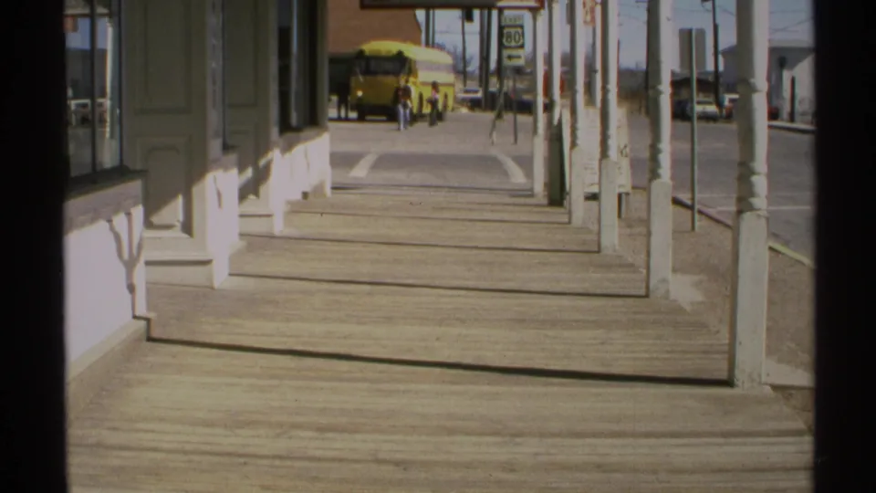 1972 Arizona 8mm frame looking down a covered boardwalk toward a yellow school bus and distant pedestrians on a small-town street.