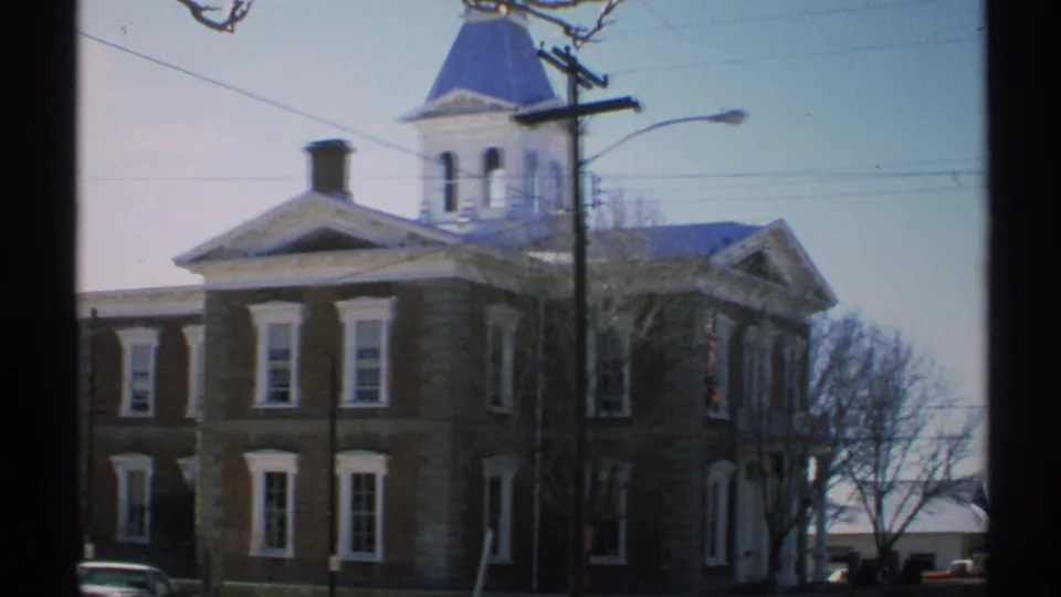 1972 Arizona 8mm frame of a large stone building with tall windows, bare trees, parked cars, and a blue steeple-like roof.
