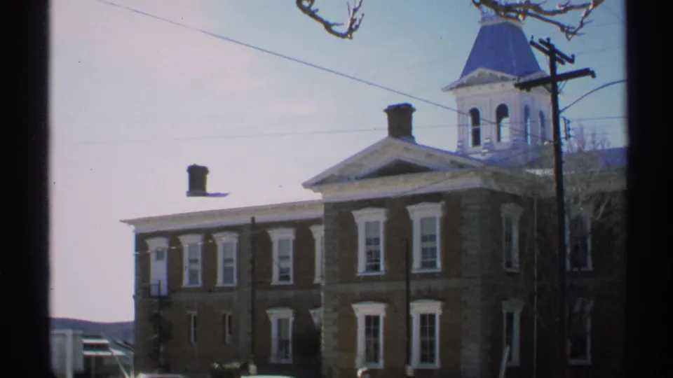 1972 Arizona 8mm frame showing more of the brick building side, white-trimmed windows, a partial cupola, and a car at the lower edge.